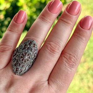 Puede incluir: Un anillo plateado adornado con una gran cara ovalada. El anillo presenta un dise&ntilde;o de filigrana detallado con peque&ntilde;os acentos oscuros. El fondo es una vista borrosa de follaje verde, y las u&ntilde;as est&aacute;n pintadas de color rosa claro.