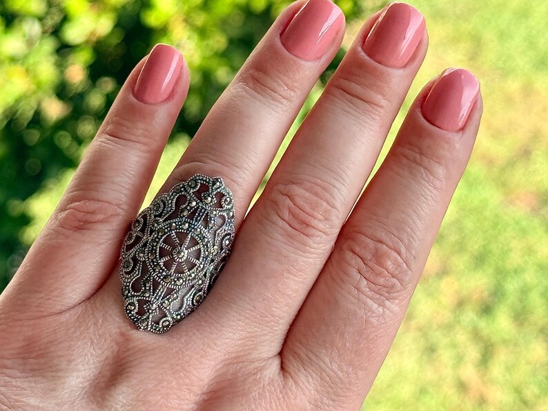 Puede incluir: Un anillo plateado adornado con una gran cara ovalada. El anillo presenta un dise&ntilde;o de filigrana detallado con peque&ntilde;os acentos oscuros. El fondo es una vista borrosa de follaje verde, y las u&ntilde;as est&aacute;n pintadas de color rosa claro.