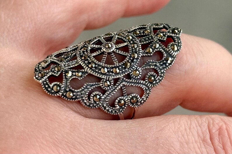 Puede incluir: Anillo plateado ornamentado con un dise&ntilde;o de filigrana. El anillo presenta un patr&oacute;n circular central y est&aacute; adornado con peque&ntilde;as piedras brillantes. Los intrincados detalles y los acentos oscuros crean una est&eacute;tica vintage.