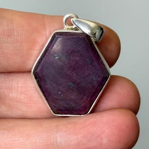 Könnte beinhalten: Ein sechseckiger Anhänger mit einem tiefvioletten Edelstein, der in einen silberfarbenen Metallrahmen gefasst ist. Der Edelstein hat eine facettierte Oberfläche und wird von einer silbernen Öse gehalten.