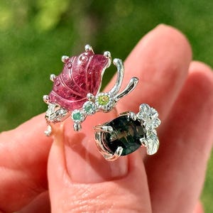 May include: A silver ring featuring a carved deep pink gemstone butterfly with small green accent stones on one side and a dark green oval gemstone with a silver floral detail on the other side, connected by a delicate silver band.