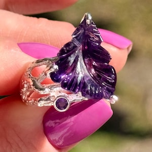 May include: A sterling silver ring featuring a carved amethyst leaf design. The ring has a detailed, organic band and a small, round amethyst accent stone. The deep purple of the amethyst contrasts with the silver.