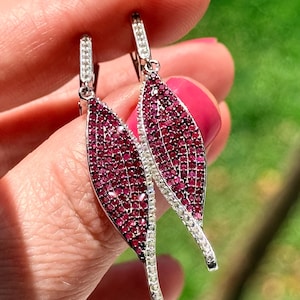 Puede incluir: Pendientes colgantes con diseño de hoja. Los pendientes presentan un engaste de metal plateado. El cuerpo principal de cada pendiente está cubierto de pequeñas piedras preciosas de color rojo intenso, con un borde de piedras claras y brillantes. Los pendientes se sujetan con un cierre plateado.