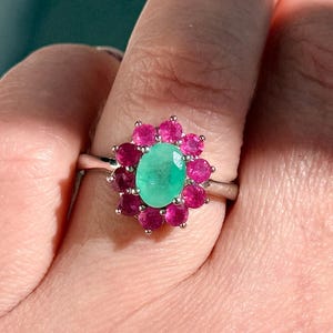 May include: A silver ring featuring a central oval-cut emerald gemstone surrounded by a halo of round, vibrant pink gemstones. The ring's design is floral, with the pink stones forming a petal-like arrangement around the green emerald.