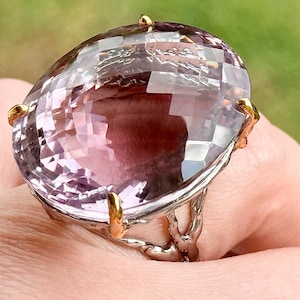May include: A large, faceted amethyst gemstone ring. The ring features a silver band with gold accents and prongs holding the large, oval-shaped purple gemstone. The ring is shown on a person's finger.