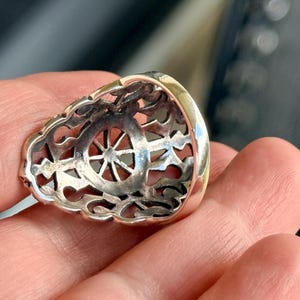 Puede incluir: Primer plano de un anillo de plata con un dise&ntilde;o calado. El anillo presenta un motivo central en forma de rueda y un borde ornamentado de estilo filigrana. El anillo se sostiene en una mano, mostrando sus detalles.