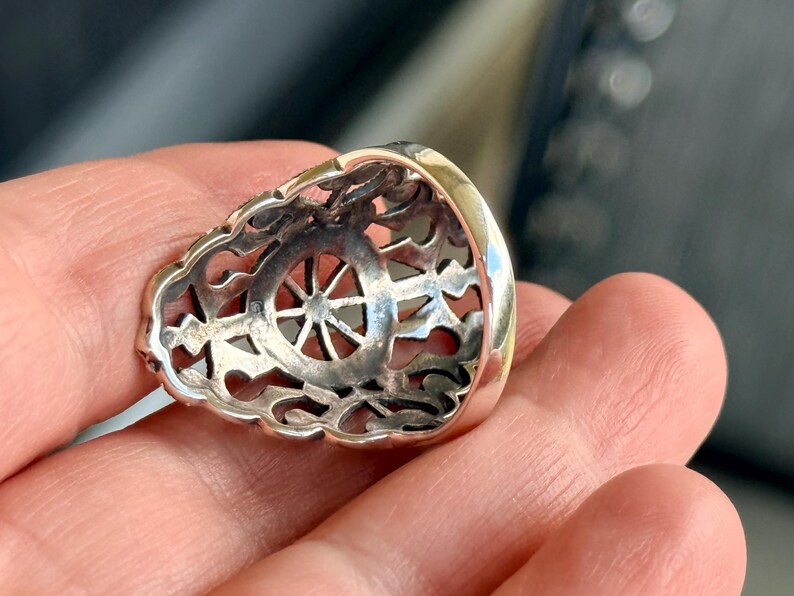 Puede incluir: Primer plano de un anillo de plata con un dise&ntilde;o calado. El anillo presenta un motivo central en forma de rueda y un borde ornamentado de estilo filigrana. El anillo se sostiene en una mano, mostrando sus detalles.