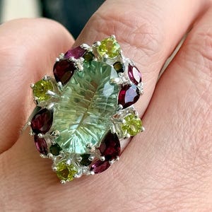 May include: A silver ring featuring a large, light green, oval-cut gemstone as the centerpiece. The ring is adorned with smaller, round and pear-shaped gemstones in shades of green and deep red. The ring is set on a finger.