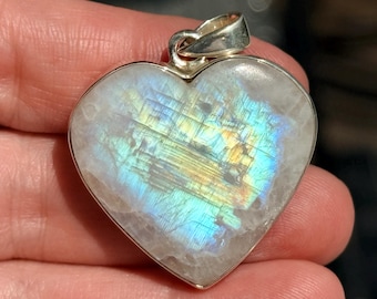 May include: Heart-shaped pendant with a silver bail. The pendant features a polished, iridescent moonstone with blue, yellow, and green hues. The stone is set in a silver bezel. A piece of jewelry.