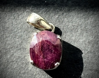 May include: A silver pendant featuring a large, oval-cut ruby gemstone. The ruby is a deep red colour with visible facets. The pendant has a simple, elegant design, with a silver bail for a chain.