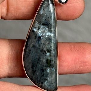 May include: A pendant with a polished, dark grey stone, featuring a silver-coloured bezel and bail. The stone has a mottled appearance with lighter grey and white inclusions, creating a unique pattern. The pendant is held between fingers.