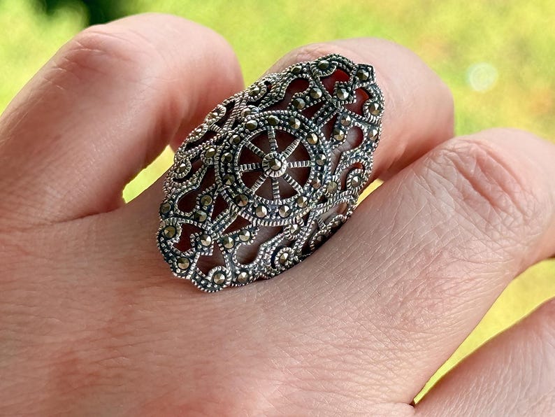 Puede incluir: Un anillo ovalado de plata con intrincados detalles de filigrana y marcasita. El anillo presenta un dise&ntilde;o central que se asemeja a una rueda o un sol, rodeado de patrones ornamentados. El anillo se lleva en un dedo.
