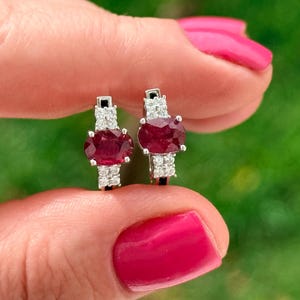 May include: A pair of silver earrings featuring oval-cut ruby gemstones, accented with smaller white gemstones. The earrings are held between fingers against a blurred green background.