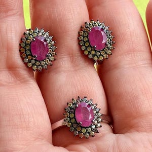 May include: A jewelry set featuring a ring and matching earrings. Each piece has a central oval ruby surrounded by small, light-colored gemstones. The ring and earrings are set in a dark metal, creating a striking contrast with the bright gemstones.