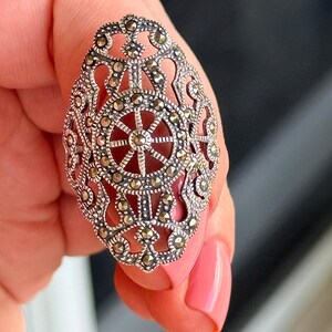 Puede incluir: Anillo ovalado de plata con intrincados detalles de filigrana y marcasita. El anillo presenta un dise&ntilde;o central con un patr&oacute;n en forma de rueda y una piedra de color marr&oacute;n rojizo. El anillo es sostenido por una persona.