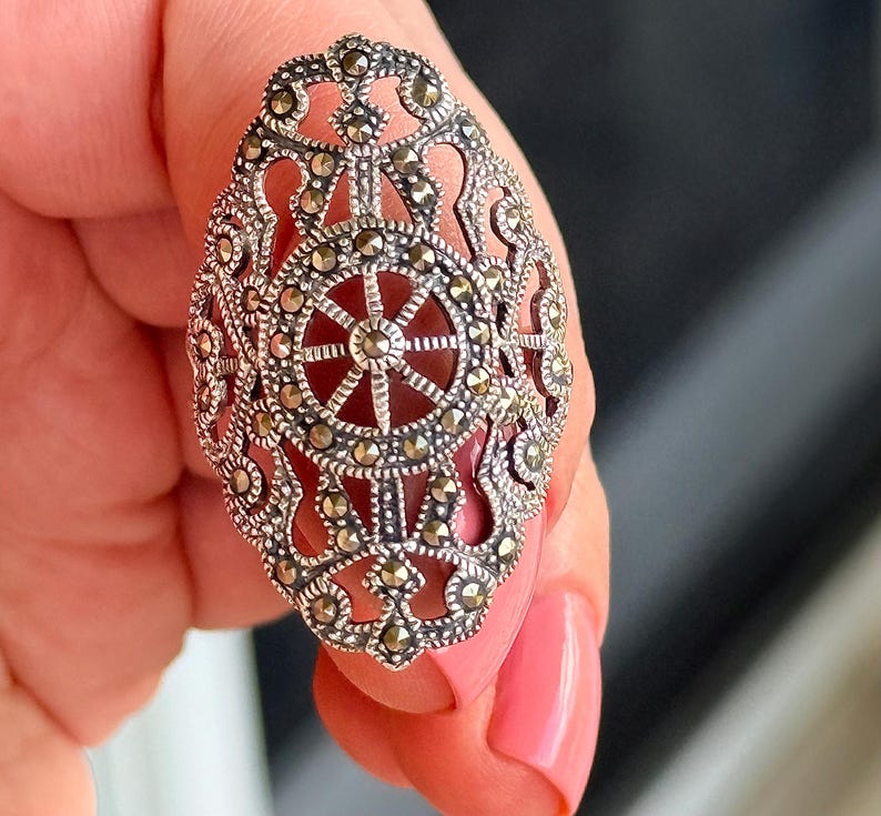 Puede incluir: Anillo ovalado de plata con intrincados detalles de filigrana y marcasita. El anillo presenta un dise&ntilde;o central con un patr&oacute;n en forma de rueda y una piedra de color marr&oacute;n rojizo. El anillo es sostenido por una persona.