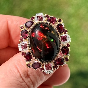 May include: An oval black opal ring with a gold and silver setting. The opal has red and green flashes. The ring is surrounded by small, round, and square red gemstones. The ring is a piece of jewelry.