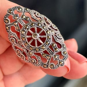 Puede incluir: Anillo plateado ornamentado con un dise&ntilde;o de filigrana y peque&ntilde;os detalles brillantes. El anillo presenta un patr&oacute;n circular central con un dise&ntilde;o de estrella. El anillo es sostenido por una persona.