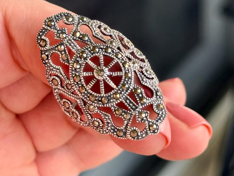 Puede incluir: Anillo plateado ornamentado con un dise&ntilde;o de filigrana y peque&ntilde;os detalles brillantes. El anillo presenta un patr&oacute;n circular central con un dise&ntilde;o de estrella. El anillo es sostenido por una persona.