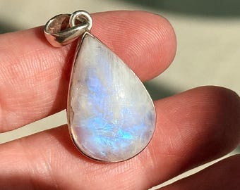 May include: A teardrop-shaped pendant with a silver bail. The pendant features a polished, iridescent moonstone with white and blue hues. The stone is set in a silver bezel. The pendant is held in a person's hand.