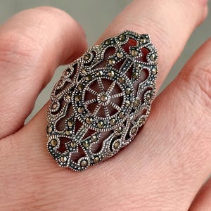 Puede incluir: Anillo de plata ovalado con un intrincado dise&ntilde;o de filigrana y un motivo central en forma de estrella. El anillo presenta peque&ntilde;as piedras brillantes. La joya se muestra en un dedo, resaltando sus detalles ornamentales.
