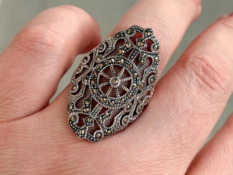 Puede incluir: Anillo de plata ovalado con un intrincado dise&ntilde;o de filigrana y un motivo central en forma de estrella. El anillo presenta peque&ntilde;as piedras brillantes. La joya se muestra en un dedo, resaltando sus detalles ornamentales.