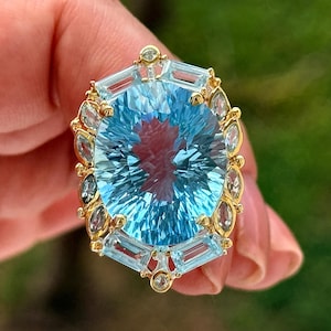 May include: A gold ring featuring a large, oval-cut, light blue gemstone at the center, surrounded by a halo of smaller, marquise-cut and baguette-cut light blue stones, with tiny round accent stones set at the top and bottom points.