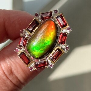 May include: A gemstone ring featuring a large, oval-shaped ammolite stone with vibrant green, orange, and yellow hues. The ammolite is surrounded by a frame of rectangular red gemstones and smaller pink stones, set in a silver and gold setting.