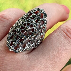 Puede incluir: Un anillo plateado ornamentado con un dise&ntilde;o de filigrana, que presenta un patr&oacute;n central en forma de estrella. El anillo est&aacute; cubierto de peque&ntilde;as piedras redondas y oscuras, y se lleva en un dedo. Los intrincados detalles del anillo se destacan sobre un fondo borroso.