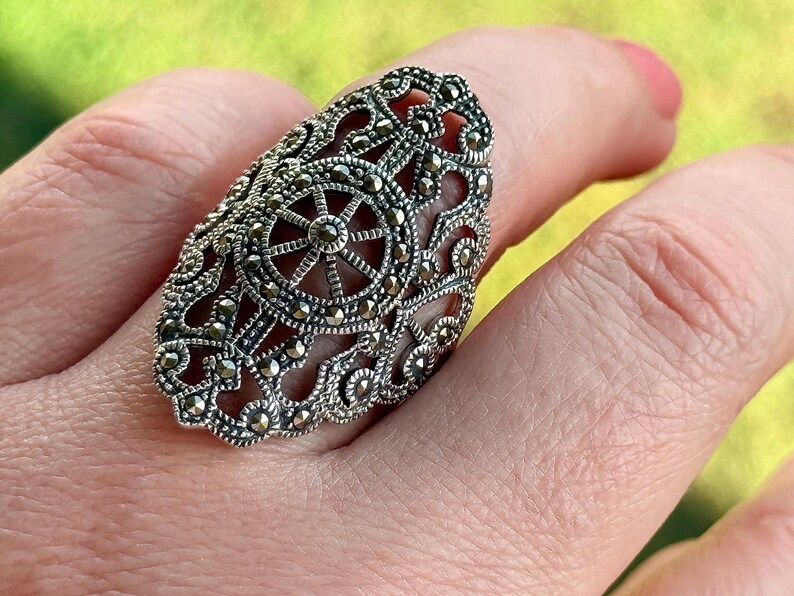 Puede incluir: Un anillo plateado ornamentado con un dise&ntilde;o de filigrana, que presenta un patr&oacute;n central en forma de estrella. El anillo est&aacute; cubierto de peque&ntilde;as piedras redondas y oscuras, y se lleva en un dedo. Los intrincados detalles del anillo se destacan sobre un fondo borroso.