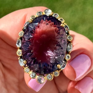 Può includere: Un grande anello ovale con una gemma sfaccettata color viola intenso come elemento centrale. La pietra centrale è circondata da un alone di pietre più piccole, rotonde, chiare e azzurre, incastonate in una montatura in metallo dorato.