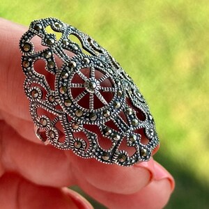 Puede incluir: Un anillo detallado, de tono plateado, con un dise&ntilde;o calado ornamentado. El anillo presenta un elemento circular central con detalles en forma de radios, rodeado de intrincados patrones y peque&ntilde;os acentos brillantes. El anillo se sostiene sobre un fondo verde borroso.