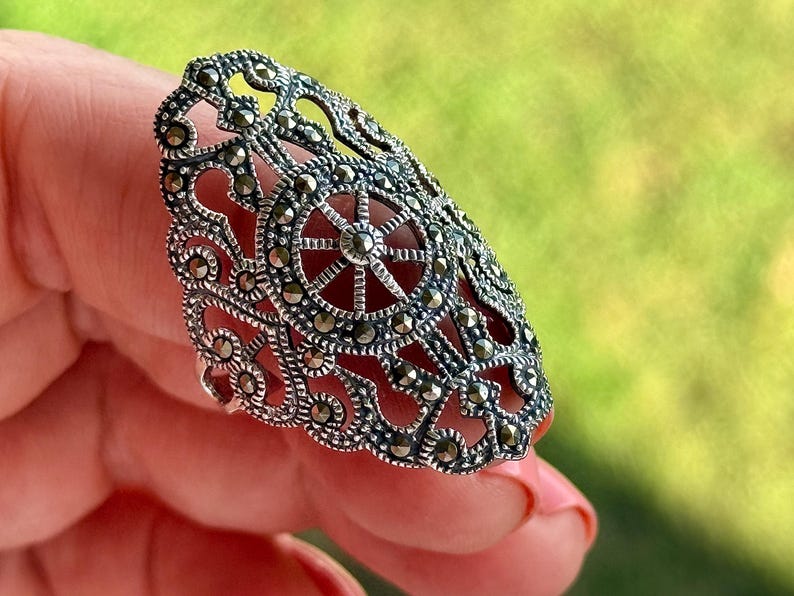 Puede incluir: Un anillo detallado, de tono plateado, con un dise&ntilde;o calado ornamentado. El anillo presenta un elemento circular central con detalles en forma de radios, rodeado de intrincados patrones y peque&ntilde;os acentos brillantes. El anillo se sostiene sobre un fondo verde borroso.