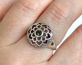 Pode incluir: Anel de prata com um design floral, apresentando uma pedra preciosa verde central e um fundo castanho-avermelhado. O anel é colocado numa banda de ouro rosa, mostrando um padrão detalhado e vazado.