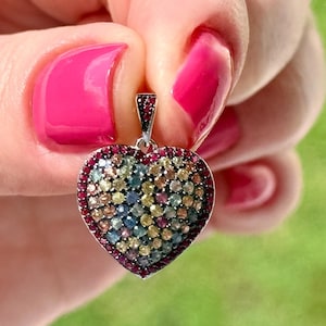 May include: A heart-shaped pendant with a silver bail. The pendant is encrusted with small, colourful gemstones in shades of red, yellow, blue, and green. The outer edge of the heart is lined with red gemstones. The background is blurred.