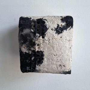 Puede incluir: Un bloque rectangular con una superficie texturizada blanquecina, parcialmente cubierta con parches negros. El bloque tiene una apariencia rugosa y envejecida, con imperfecciones visibles y pequeños agujeros. La estética general sugiere un artículo desgastado o artesanal.