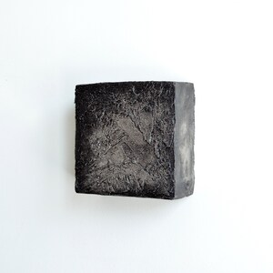 May include: A small, square, black sculpture with a textured surface. The sculpture has a rough, uneven texture and appears to be made of a dark, heavy material.