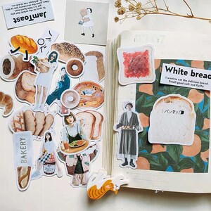 Peut inclure: Une collection d'autocollants colorés représentant différents types de pain, notamment des miches, des bagels et des toasts. Les autocollants comprennent également des illustrations de personnes et d'un chat. Le texte "Pain blanc" est écrit sur l'un des autocollants, ainsi que la phrase "J'ai envie de manger ce délicieux pain. Il sent bon, il est moelleux et doux."