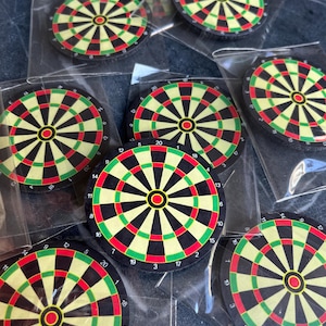 May include: Multiple small, circular dartboard designs, each in a clear plastic packet. The dartboards have a classic colour scheme of black, red, and green, with numbers around the edge. The bullseye is red.