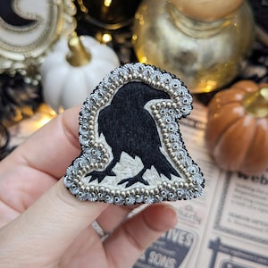May include: A detailed raven brooch featuring a black bird silhouette, embellished with silver and gold beads. The raven is set on a white background. A unique accessory for Halloween or gothic-themed outfits.