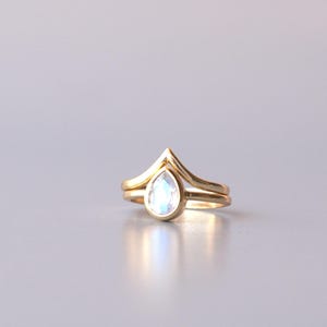 May include: A gold ring set consisting of two bands. One band features a pear-shaped moonstone with a blue iridescent sheen, set in a bezel. The second band is a V-shaped chevron band that nests against the stone setting.