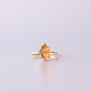 May include: A gold ring featuring a single, faceted, kite-shaped orange gemstone held in place by four delicate prongs. The band is thin and polished, showcasing a simple design that highlights the geometric shape and warm color of the stone.