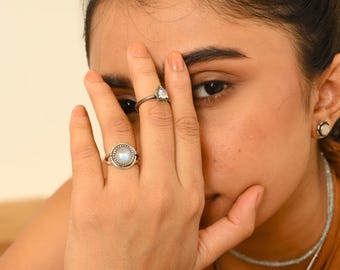 May include: Two silver rings featuring iridescent moonstone gems. One ring has a large, round, faceted moonstone in a serrated setting, while the other features a smaller, pear-shaped moonstone. Both rings are worn on the fingers of a hand.