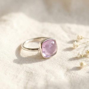 May include: A silver ring featuring a smooth, cushion-cut light purple gemstone set in a simple metal band. The gemstone has a translucent appearance and a polished surface, reflecting light across its rounded, square-shaped top profile.