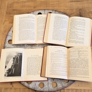 May include: Four open books with different titles and text. The books are on a wooden table with a metal spool in the center. The books are all open to different pages and show different text. The books are all in good condition and have no visible damage.