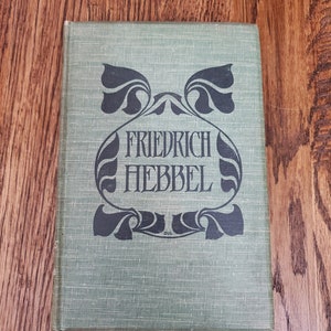 Pu&ograve; includere: Un libro rilegato in verde con un designo decorativo in bianco e nero sulla copertina. Il titolo "FRIEDRICH HEBBEL" &egrave; stampato in lettere nere.