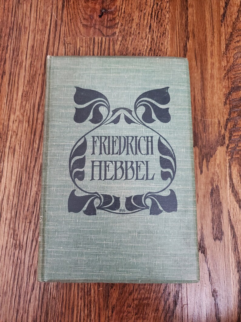 Pu&ograve; includere: Un libro rilegato in verde con un designo decorativo in bianco e nero sulla copertina. Il titolo "FRIEDRICH HEBBEL" &egrave; stampato in lettere nere.