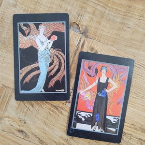 May include: Two vintage cards with black borders featuring women in elegant dresses and a phoenix bird. The woman on the left card is holding a red apple. The woman on the right card is wearing a black dress with blue flowers.