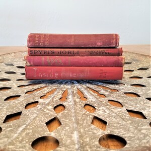 May include: A stack of four red vintage hardcover books with the titles "First Greek Book", "Spyris Jorli", "Clayton Wunderli", and "Inside Baseball". The books are sitting on a round, metal, decorative tray with a white painted finish and a cut-out design.