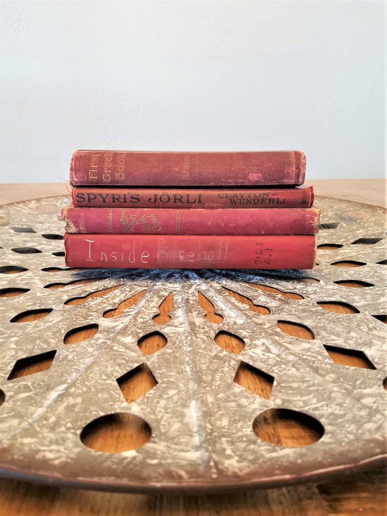 May include: A stack of four red vintage hardcover books with the titles "First Greek Book", "Spyris Jorli", "Clayton Wunderli", and "Inside Baseball". The books are sitting on a round, metal, decorative tray with a white painted finish and a cut-out design.